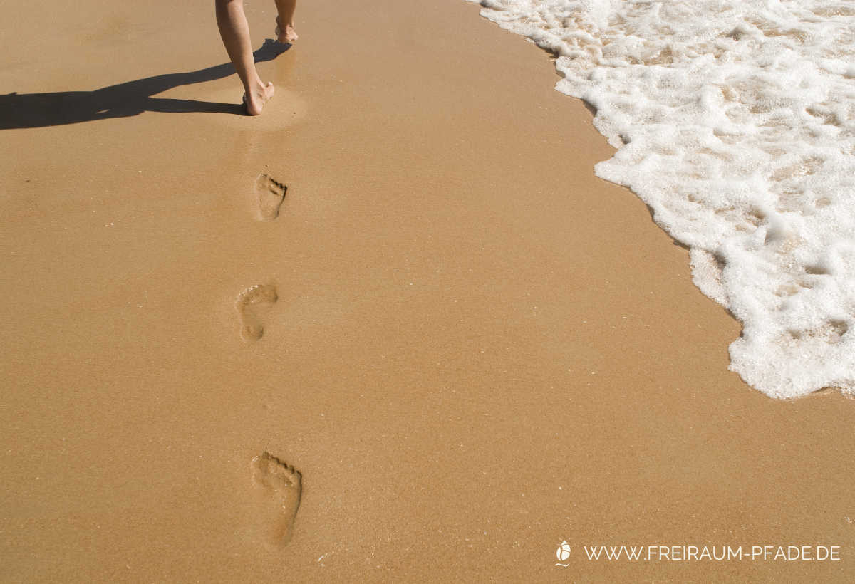 Von hinten sieht man zwei nackte Unterschenkel, die im rötlich-braunen Sand am Meer Fußabdrücke hinterlassen – rechts ist weißer Schaum der Meeresbrandung zu sehen.