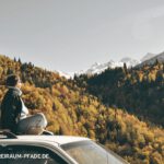 Person sitzt ruhig auf einem Autodach mit Blick auf bunte Herbstwälder und schneebedeckte Berge – Symbol für innehalten, Naturverbindung und innere Weite