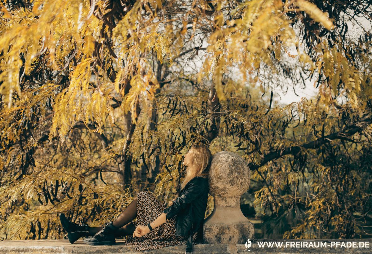 Frau sitzt an einem Steinpfeiler unter herbstlich goldenen Bäumen – Sinnbild für Natur im Alltag erleben in nur 10 Minuten