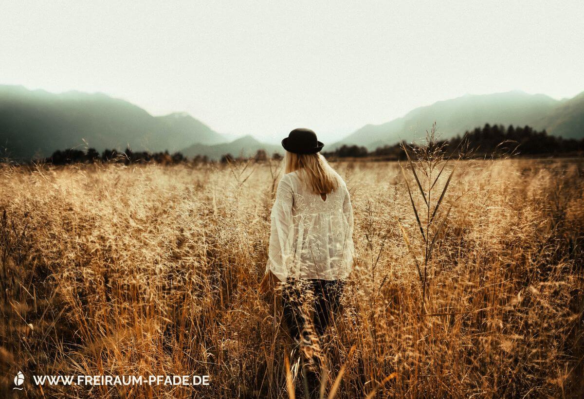Frau mit Hut steht allein in goldenem Grasfeld vor Bergkulisse - symbolisiert die Suche nach eigenem Raum und eigenen Pfaden in der Natur