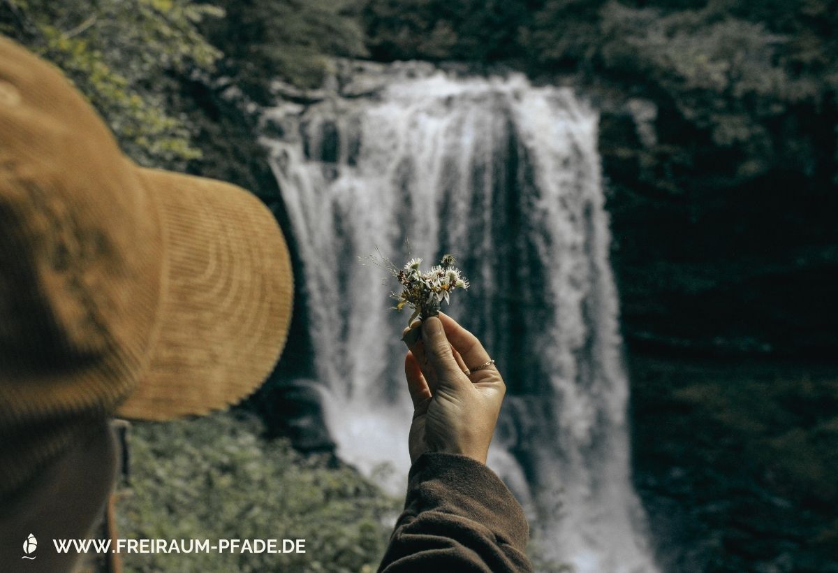 Eine Frau mit braunem Sonnenhut hält eine weiße Blüte in der Hand, im Hintergrund vor einem Wasserfall