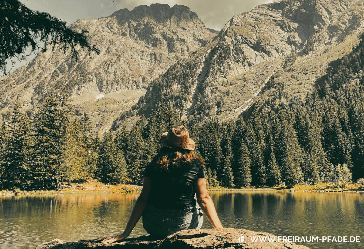 eine Frau mit roten Haaren und Strohhut sitzt auf einem Felsen an einem See mit Tannenbäumen am Ufer vor einer Bergkulisse