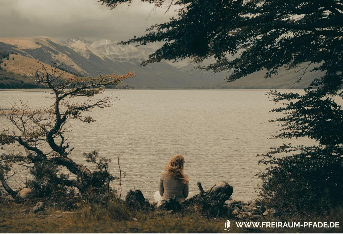 eine Frau mit wehenden blonden Haaren sitzt entspannt unter einem Baum am Ufer eines Bergsees